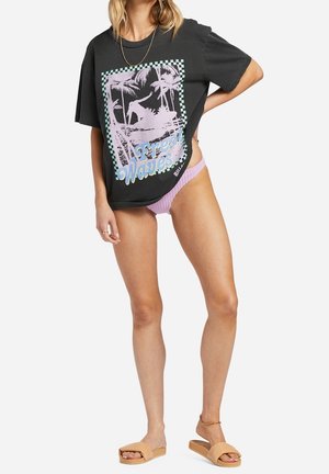 Frau trägt übergroßes schwarzes T-Shirt mit pastellfarbenem Surfmotiv, lavendelfarbene Bikinihose und braune Sandalen, steht mit der Hand auf der Hüfte.