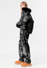 Chaqueta negra con patrón y cuello de piel sintética, combinada con pantalones anchos a juego. La modelo lleva botas de plataforma naranjas para crear contraste.