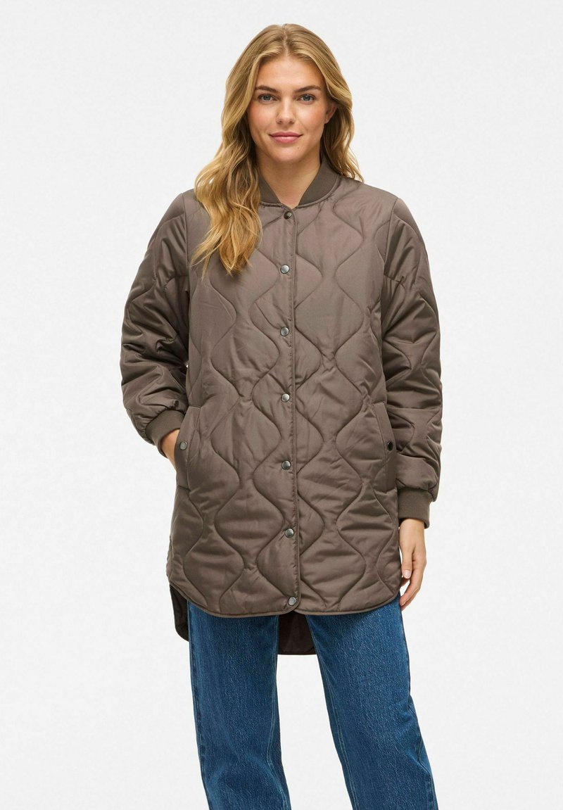 Vrouw met lang blond haar, gekleed in een bruine gewatteerde button-up jas en blauwe jeans, staand met haar handen in de jaszakken.