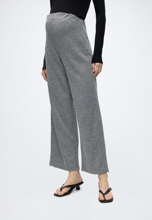 Personne porte un pantalon gris taille haute à jambes larges, un haut noir à manches longues et des sandales noires à talons sur fond blanc.