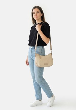 Femme portant un chemisier noir et un jean bleu clair, tenant un sac à bandoulière beige, debout avec des baskets blanches devant un fond uni.