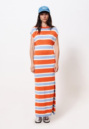 Gestreepte gebreide maxi-jurk in oranje, lichtblauw en crème, met korte mouwen en een zijsplit. Gecombineerd met zilveren schoenen en een blauwe pet.