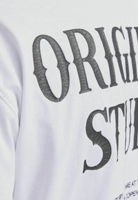 Camiseta blanca de algodón con grandes letras en relieve de color negro que dicen "ORIGINAL STUD", con una superficie de tela suave y lisa.