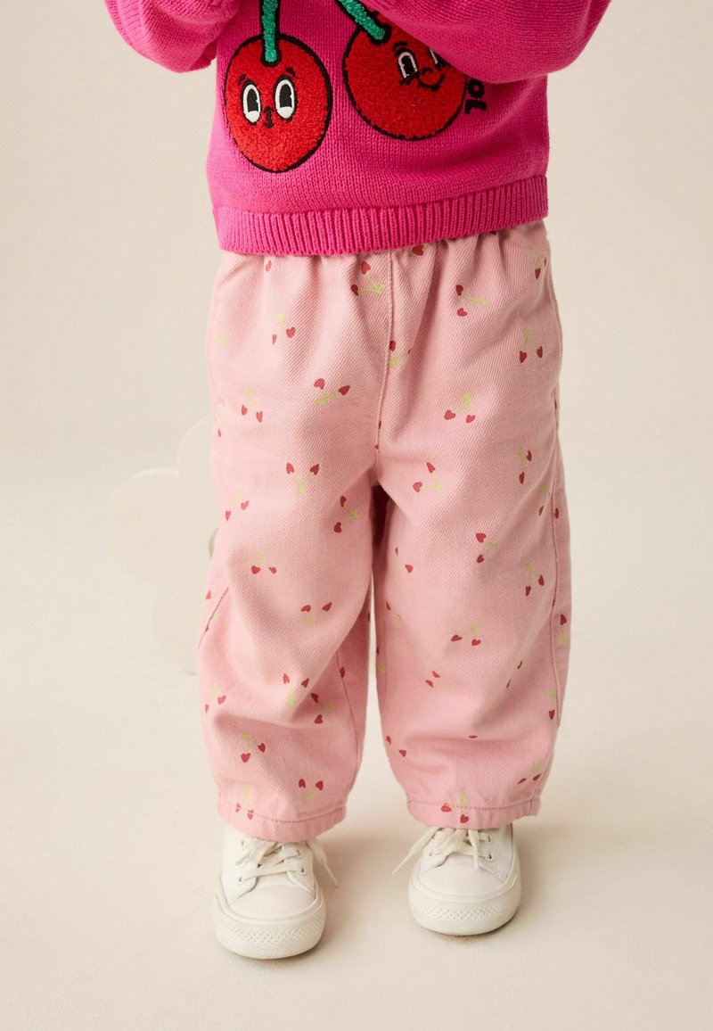 Pantaloni in corduroy rosa con una vestibilità rilassata, caratterizzati da un motivo di piccoli cuori rossi e steli verdi. Indossati con sneakers bianche.