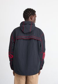 Chaqueta negra con costuras y acentos rojos, que cuenta con capucha, mangas largas y un corte relajado. Detalle notable del logo en la manga izquierda.