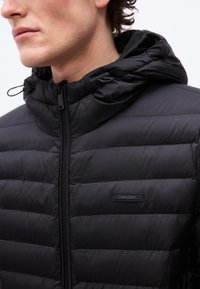 Κοντινό πλάνο ενός άνδρα που φοράει μαύρο καπιτονέ μπουφάν Calvin Klein με φερμουάρ μπροστά και κουκούλα, εμφανίζοντας το πλαϊνό προφίλ του προσώπου του.