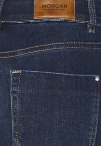 Jeans de mezclilla azul oscuro con una etiqueta de cuero que dice "MORGAN." Presenta costuras doradas contrastantes y un bolsillo con un detalle de remache metálico.