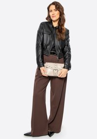 Giubbotto di pelle nera cropped, aderente in vita, abbinato a pantaloni larghi marroni. Tiene in mano una clutch beige con borchie e dettagli con fibbia.