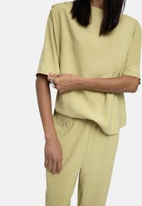 Jasnozielona bluzka z krótkim rękawem w luźnym kroju, wykonana z miękkiego materiału, z okrągłym dekoltem i dekoracyjnymi guzikami na ramieniu.