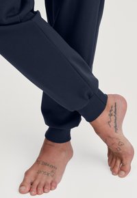 Marinefarbene Jogginghosen mit elastischen Bündchen und glatter Textur, kombiniert mit barfüßigen Füßen, die verschiedene Tattoos zeigen.