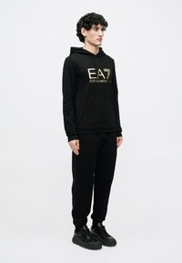 Mladý muž stojí, oblečený v čiernej mikine s kapucňou so zlatým logom "EA7 Emporio Armani", čiernych teplákoch a čiernych teniskách na bielom pozadí.