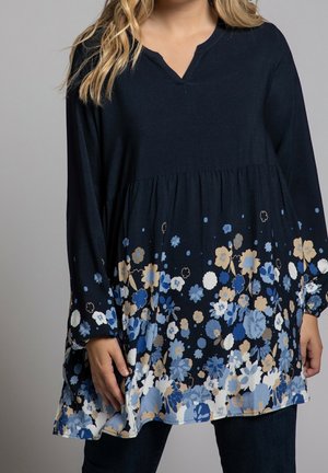 Femme portant une tunique bleu marine à manches longues avec des motifs floraux bleus, beiges et blancs sur la moitié inférieure et les manches.