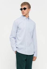 Blau-weiß gestreiftes Langarmhemd mit Knopfverschluss und Tasche. Trägt man mit grünen Hosen und ovalen Sonnenbrillen. Glatte Textur, kragenförmiges Design.