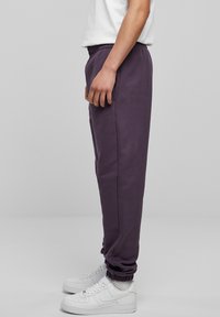 Lila sweatpants med elastisk midja, sidfickor och avskurna anklar. Bärs med vita sneakers, på en enkel grå bakgrund.