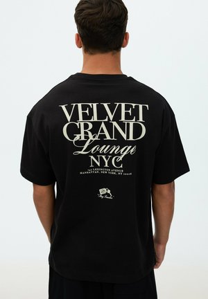 Hombre con camiseta negra con texto blanco que dice "Velvet Grand Lounge NYC, 147 Lexington Avenue, Manhattan, New York, NY 10016" en la espalda.