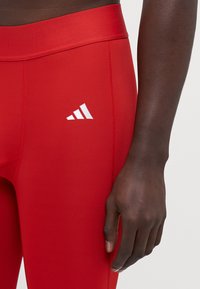 Røde atletiske leggings med en bred elastisk talje, glat stoftekstur og et hvidt Adidas-logo på venstre lår.