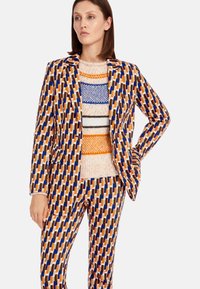 Muster-Blazer und Hosen-Set in Orange, Blau und Braun mit geometrischen Formen. Darunter ein pastellfarbener Pullover mit gestreiften Akzenten.