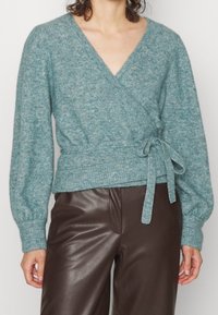 Teal omslags-tröja i mjukt stickat material, med långa puffärmar, djup V-ringning och knytbar midja, kombinerad med bruna läderbyxor.