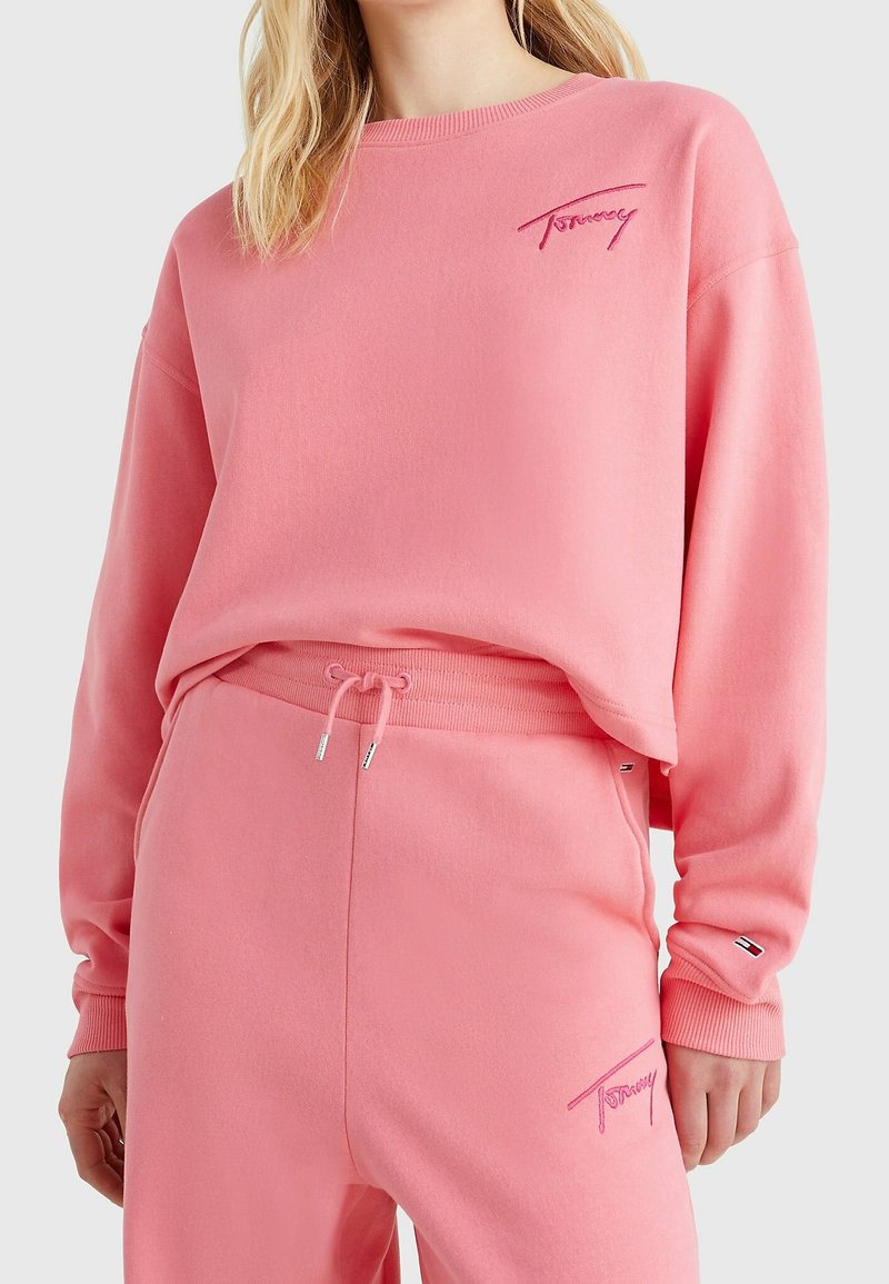 Femme portant un sweat-shirt et un pantalon de survêtement roses assortis avec "Tommy" brodé sur la poitrine et la cuisse, debout devant un fond uni.