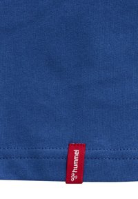 Blåt stof i close-up med et lille rødt tøjmærke med hvid "hummel" tekst og logo nær kanten.