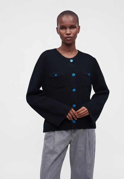 Femme portant un cardigan noir avec des boutons bleus et des poches poitrine, associé à un pantalon gris ample, debout devant un fond blanc uni.