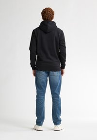Felpa nera in cotone con tasca posteriore, abbinata a jeans blu chiaro e sneakers bianche, caratterizzata da una texture liscia e cappuccio con cordini regolabili.