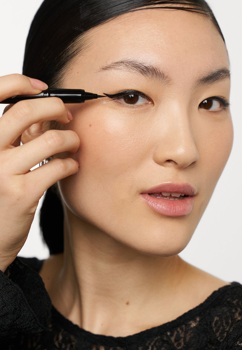 Eyeliner nero a pennarello applicato sulla palpebra superiore da una persona. La pelle è liscia, con una leggera luminosità; lo sfondo è neutro.