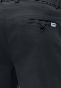 Nahaufnahme schwarzer, maßgeschneiderter Hosen mit einer geknöpften Gesäßtasche und einem kleinen weißen Lindbergh Markenetikett.