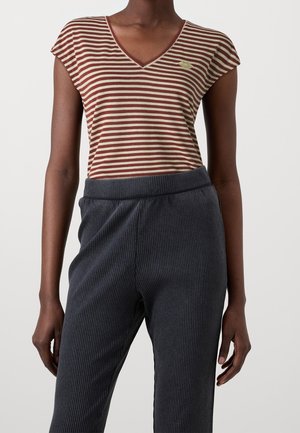 Persoon draagt een korte mouw, bruin-wit gestreept V-hals shirt met een klein logo op de borst en donkere geribbelde broek.