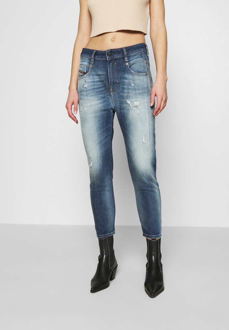 Slim-fit blue denim jeans with distressed details, mid-rise waist, and cuffed ankles. Worn with black ankle boots and a beige crop top.