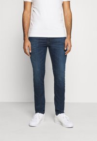 Jeans in denim slim fit di colore blu scuro con una texture liscia. Indossati con una t-shirt bianca semplice e sneakers bianche. Design classico a cinque tasche.