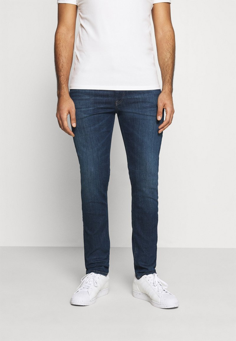 Jeans in denim slim fit di colore blu scuro con una texture liscia. Indossati con una t-shirt bianca semplice e sneakers bianche. Design classico a cinque tasche.