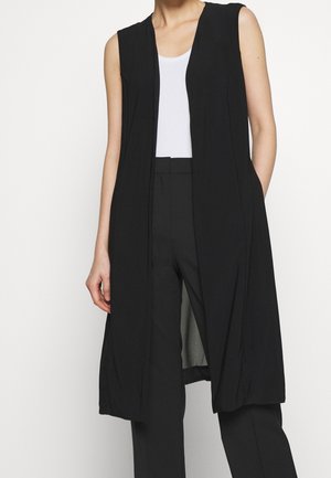 Vrouw draagt een mouwloos lang zwart vest over een witte top en zwarte broek, staand tegen een effen lichte achtergrond.