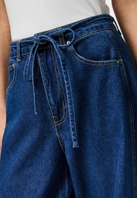 Tamno plave traperice s remenom u struku, s petljama za remen, prednjim džepovima i žutim šavovima. Glatka tekstura i opušteno krojenje.