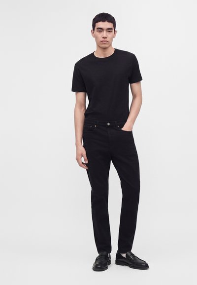 Jeune homme debout avec une main dans la poche, portant un t-shirt noir, un pantalon noir et des chaussures noires sans lacets, sur un fond clair uni.