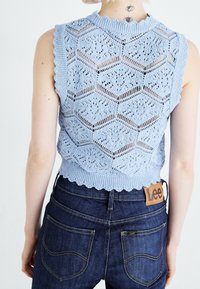 Ljusblått stickat ärmlöst topp med ett hexagonalt spetsmönster och vågiga kanter, parat med mörka jeanstyg med synliga sömmar.