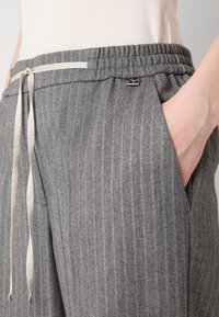 Personne portant un pantalon gris texturé avec une ceinture élastique et un cordon clair, main reposant dans la poche latérale.