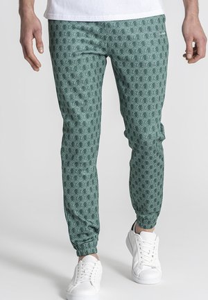 Hombre que lleva pantalones verdes ajustados con un patrón geométrico blanco repetido y zapatillas blancas, de pie sobre un fondo claro.
