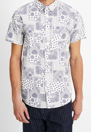 Chemise blanche à manches courtes avec un motif patchwork de cachemire bleu et géométrique, portée avec un pantalon sombre à fines rayures.