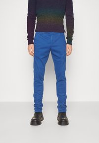 Siniset puuvillahousut, joissa on slim fit -leikkaus, litteä etuosa ja yksityiskohtaiset tikkaukset, yhdistettynä mustiin korkokenkiin ja moniväriseen neulepaitaan.