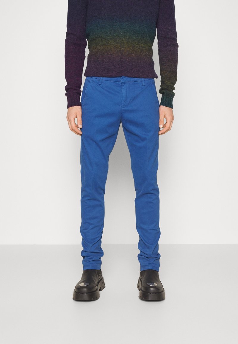 Siniset puuvillahousut, joissa on slim fit -leikkaus, litteä etuosa ja yksityiskohtaiset tikkaukset, yhdistettynä mustiin korkokenkiin ja moniväriseen neulepaitaan.