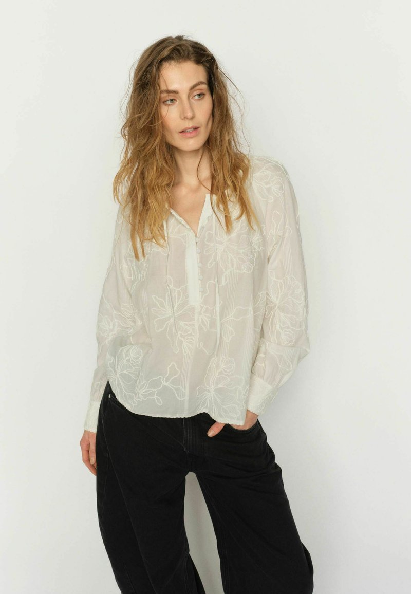 Laza, hullámos hajú nő, fehér, virágos hímzéses blúzt és fekete nadrágot visel, egyik kezét zsebében tartva, egyszínű háttér előtt áll.