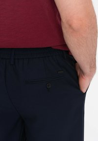Pantaloni navy con vita elastica, frontale liscio e tasche laterali. Dettagli notevoli includono una piccola tasca con chiusura a bottone e un tessuto testurizzato.