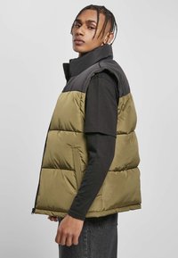 Puffervest med sort overdel og olivengrøn nederste sektion, med høj krave, lynlås og sidelommer; glat tekstur.