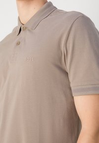Homme portant un polo marron clair avec deux boutons et "BOSS" brodé sur la poitrine, visible du cou au milieu du torse.