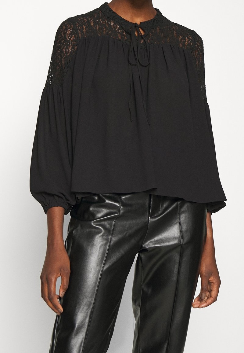 Blouse noire avec détail en dentelle sur les épaules, dotée d'un col rond et d'une fermeture à nouer, associée à un pantalon noir brillant en simili cuir.