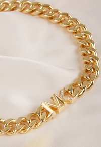 Collana in oro con maglie grasse e un accento con logo decorato con gemme trasparenti, su uno sfondo beige morbido.