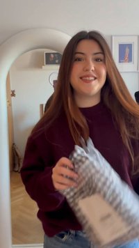 Mladá žena sa usmieva a drží darček zabalený v károvanom papieri v miestnosti s veľkým oblúkovým zrkadlom a obrazmi v rámikoch na stene.