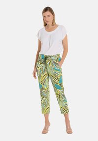 Weißes kurzärmliges Oberteil, kombiniert mit lebhaft grünen und gelben gemusterten Hosen, die Blattdesigns und geometrische Akzente aufweisen. Transparente Sandalen mit Knöchelriemen.