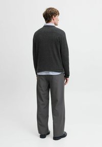 Homme portant un pull gris foncé, une chemise bleue clair, un large pantalon gris et des chaussures noires, se tenant de dos devant un fond uni.